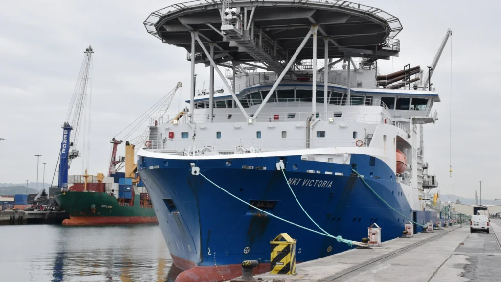 Proa del buque cablero NKT Victoria, de casco azul y superestructura blanca con helipuerto, amarrado en un muelle del puerto de Bilbao junto a un carguero y grúas de contenedores