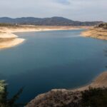 Embalse de Entrepeñas, en Guadalajara, con el nivel del agua bajo y amplias orillas de tierra clara al descubierto, rodeado de pinos y montes bajo un cielo nublado.